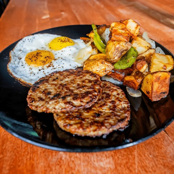 A Sunrise Social breakfast plate with two fried eggs, two sausage patties, and roasted potatoes with peppers.
