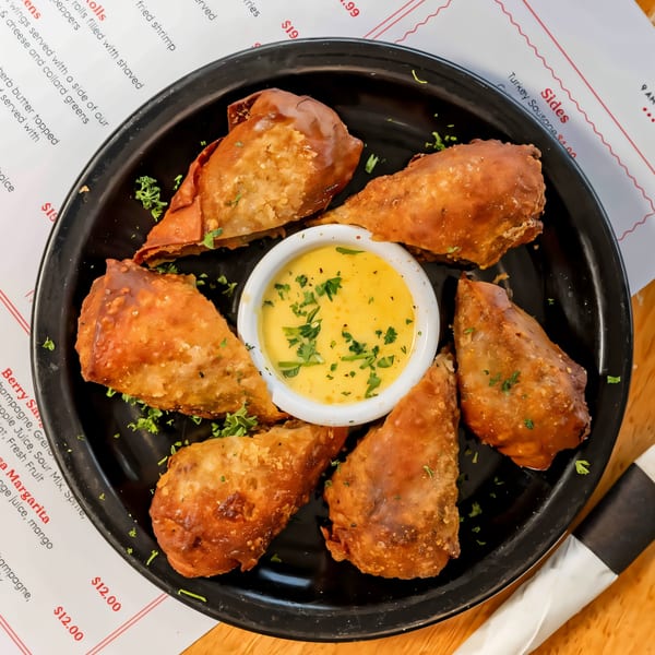 Six crispy, golden-brown breakfast egg rolls with a side of creamy dipping sauce on a black plate.
