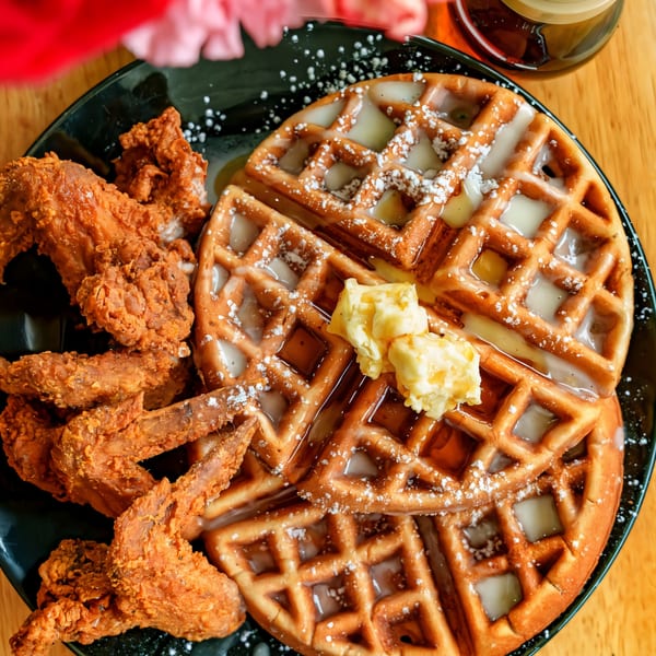 Golden-brown chicken and waffles with syrup, powdered sugar, and butter on a black plate.