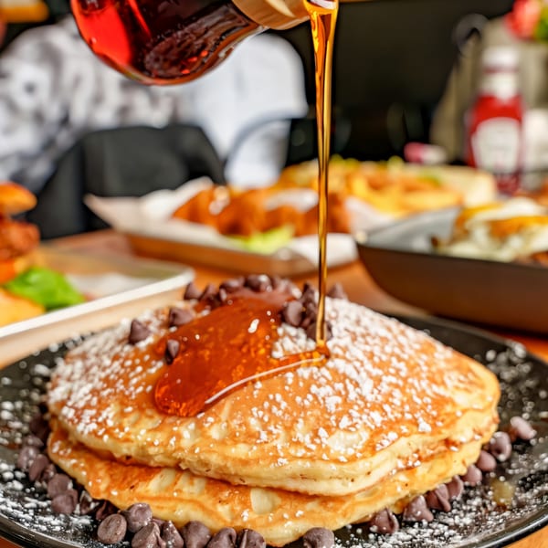 Maple syrup pouring over chocolate chip pancakes dusted with powdered sugar at Sunrise Social.