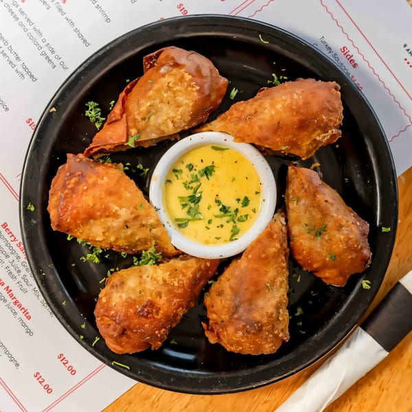Six crispy fried wontons with a side of yellow dipping sauce, garnished with parsley on a black plate.