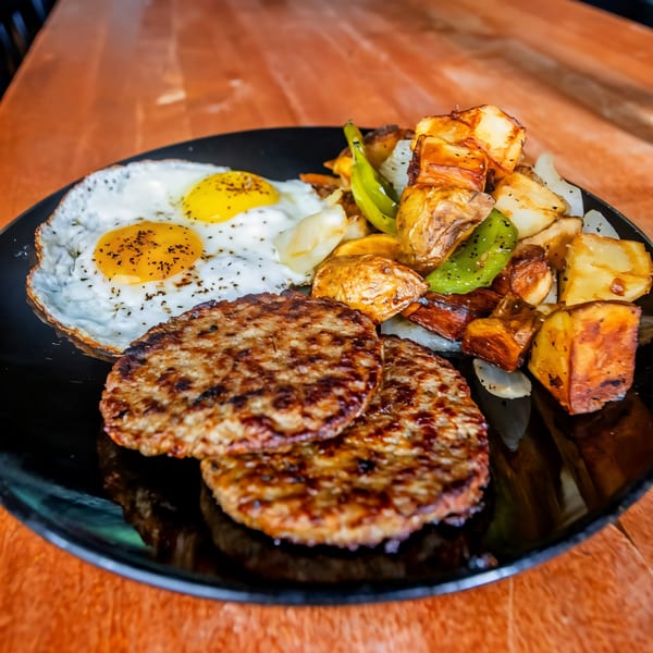 A Sunrise Social breakfast plate with two fried eggs, two sausage patties, and roasted potatoes with peppers.